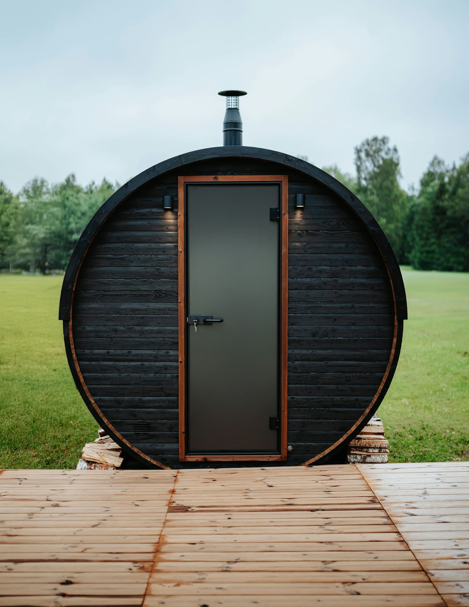 Black barrel sauna on a wood deck set in a green backyard lawn